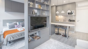 Una habitación moderna con cama, televisor, estanterías empotradas, pequeña cocina y mesa de comedor.