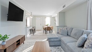 Een woonkamer met een bank, een salontafel, een aan de muur bevestigde televisie, een eethoek met een tafel en stoelen, en een potplant.