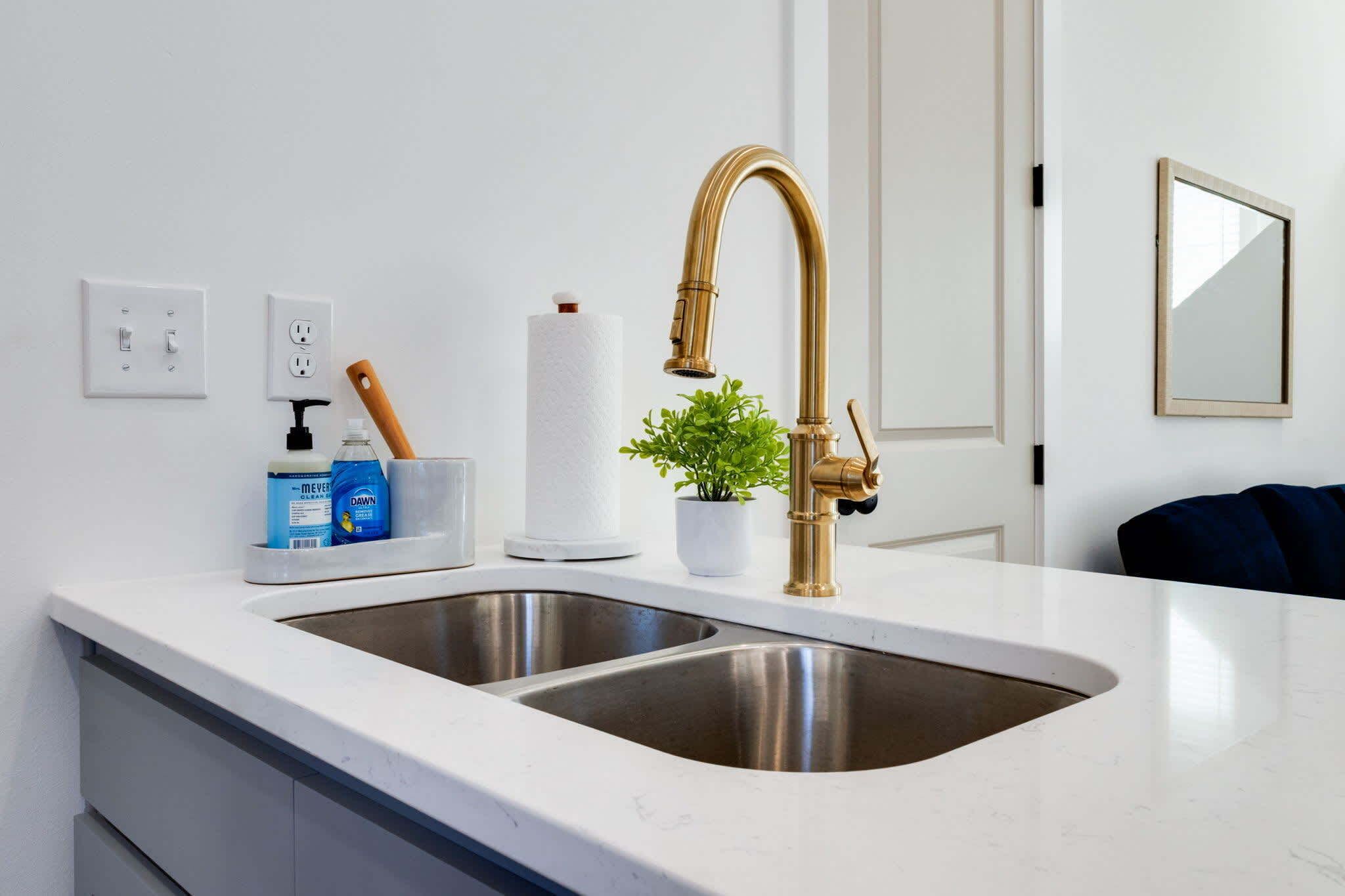 A kitchen with a double sink, a gold faucet, a potted plant, and a white countertop.