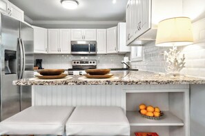 Cocina moderna con gabinetes blancos, electrodomésticos de acero inoxidable, encimeras de granito y un cuenco de naranjas en un estante.