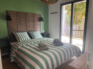 Un dormitorio con cama, ropa de cama verde a rayas, cabecera de madera, mesita de noche, lámpara y ventana con persianas.