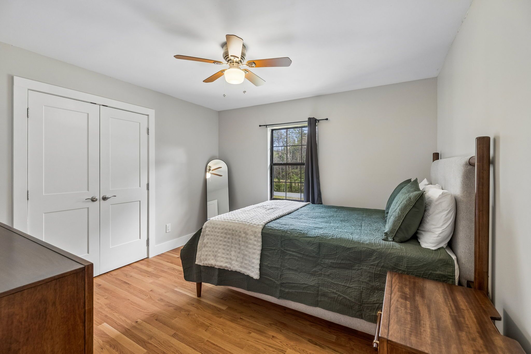 Une chambre à coucher comprenant un lit, un ventilateur de plafond, une fenêtre avec des rideaux, une porte et une commode en bois.