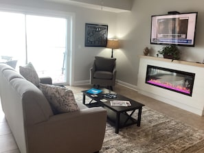 Ein Wohnzimmer mit Sofa, Sessel, Couchtisch, Kamin, Fernseher und einer Tür, die auf einen Balkon führt.