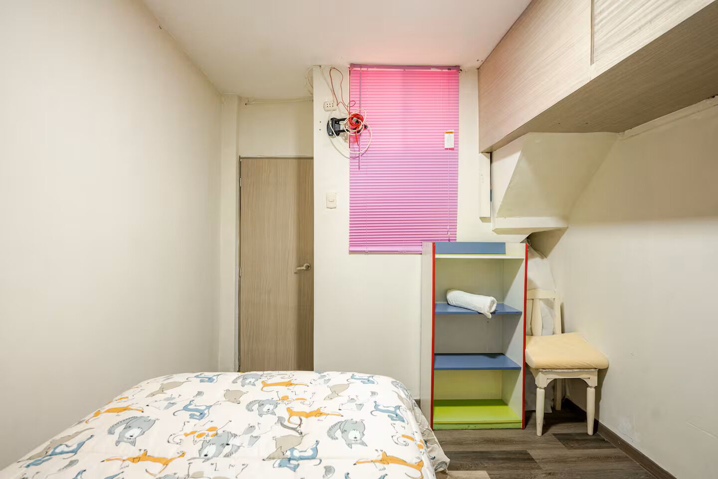 A bedroom with a bed, a window with blinds, a shelf unit, and a chair.