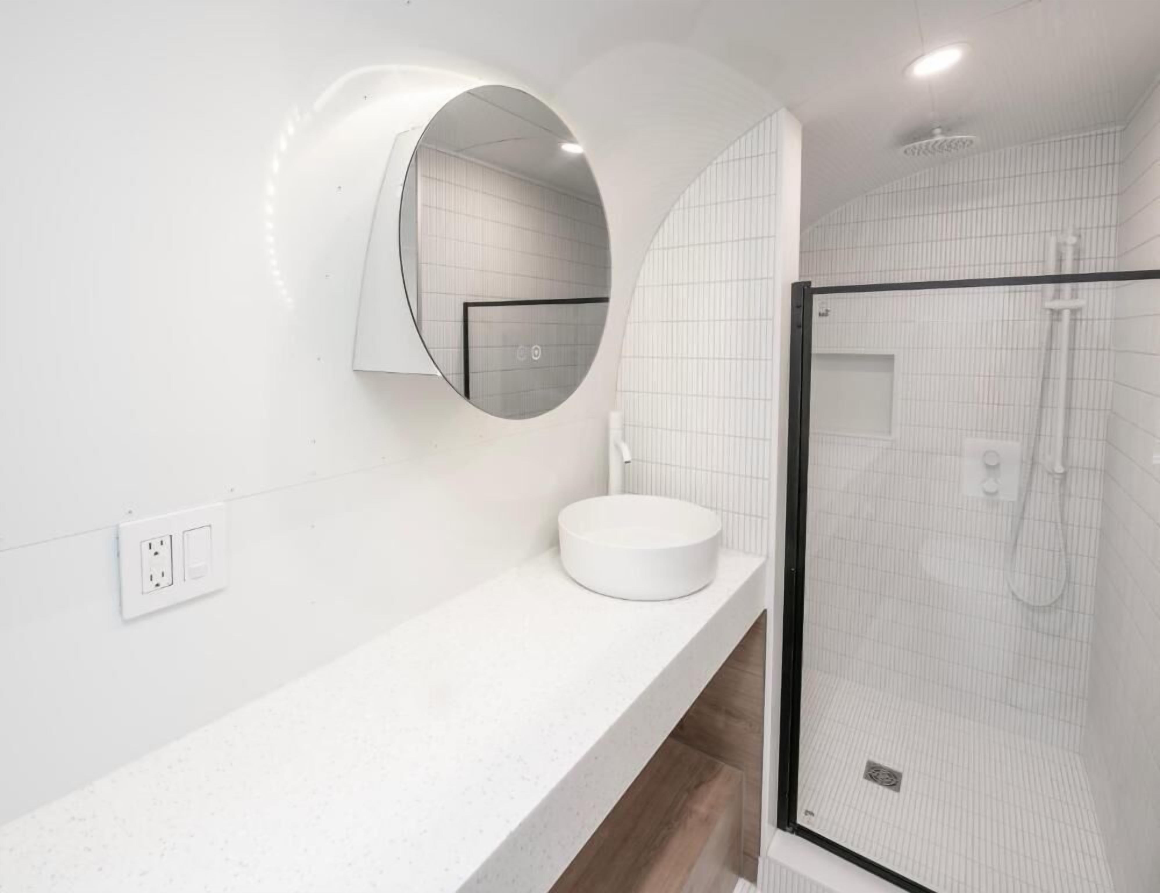 Un baño moderno con un espejo redondo, un lavabo blanco y una ducha con mampara de vidrio.
