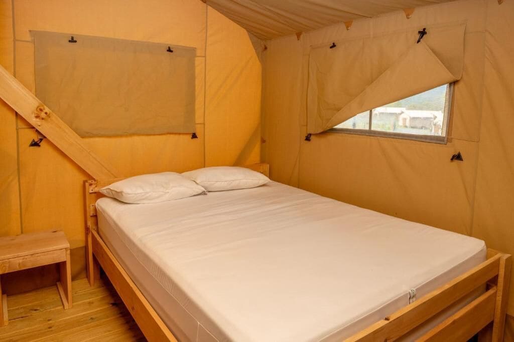 Un lit simple avec un cadre en bois et du linge de lit blanc, un petit banc en bois et une fenêtre donnant sur les bâtiments à l’extérieur.