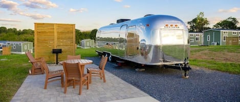 A silver trailer with 'AIRSTREAM' branding, a wooden deck with chairs and a table, and a small shed in the background.