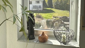 Una habitación con ventana que da a una casa y un jardín, un estante pequeño con un jarrón y un cuenco decorativo, y una planta a la izquierda.