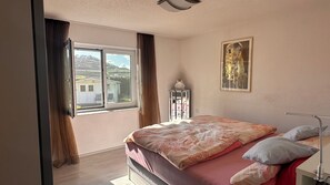 Ein Schlafzimmer mit großem Fenster, einem Bett mit gemusterten Textilien, einem Nachttisch mit Lampe und einem gerahmten Kunstwerk an der Wand.