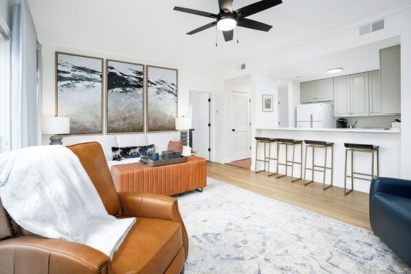 A modern living room with a brown leather sofa, a blue armchair, and a white rug. There are three bar stools and a white refrigerator in the kitchen area.