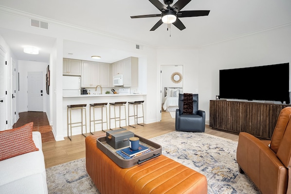 A modern living room with a sofa, ottoman, and a coffee table with a tray. There is a TV, a ceiling fan, and a kitchen area in the background.