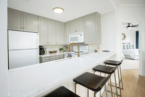 A modern kitchen with white cabinetry, a white countertop, a built-in oven, and a microwave. There are four stools at the counter.