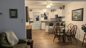 Ein Wohnzimmer mit Sofa, einem Esstisch mit Stühlen, einer Küchenzeile inklusive Schränke und Geräte sowie einem Deckenventilator.