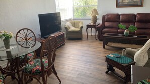 Ein Wohnzimmer mit Fernseher, einer Couch, einem Esstisch mit Stühlen und einem Fenster mit Jalousien.