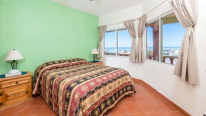 Ein Schlafzimmer mit Bett, Nachttisch, Lampe, Deckenventilator, einem Fenster mit Vorhängen und Blick auf das Meer.