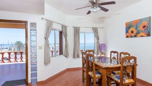 Un comedor con una mesa de vidrio, sillas de madera, un ventilador de techo y vista al mar.