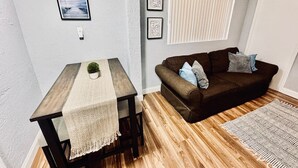 Ein Zimmer mit Holzboden, einem braunen Sofa mit Kissen, einem kleinen Tisch mit weißer Tischdecke und gerahmten Bildern an der Wand.