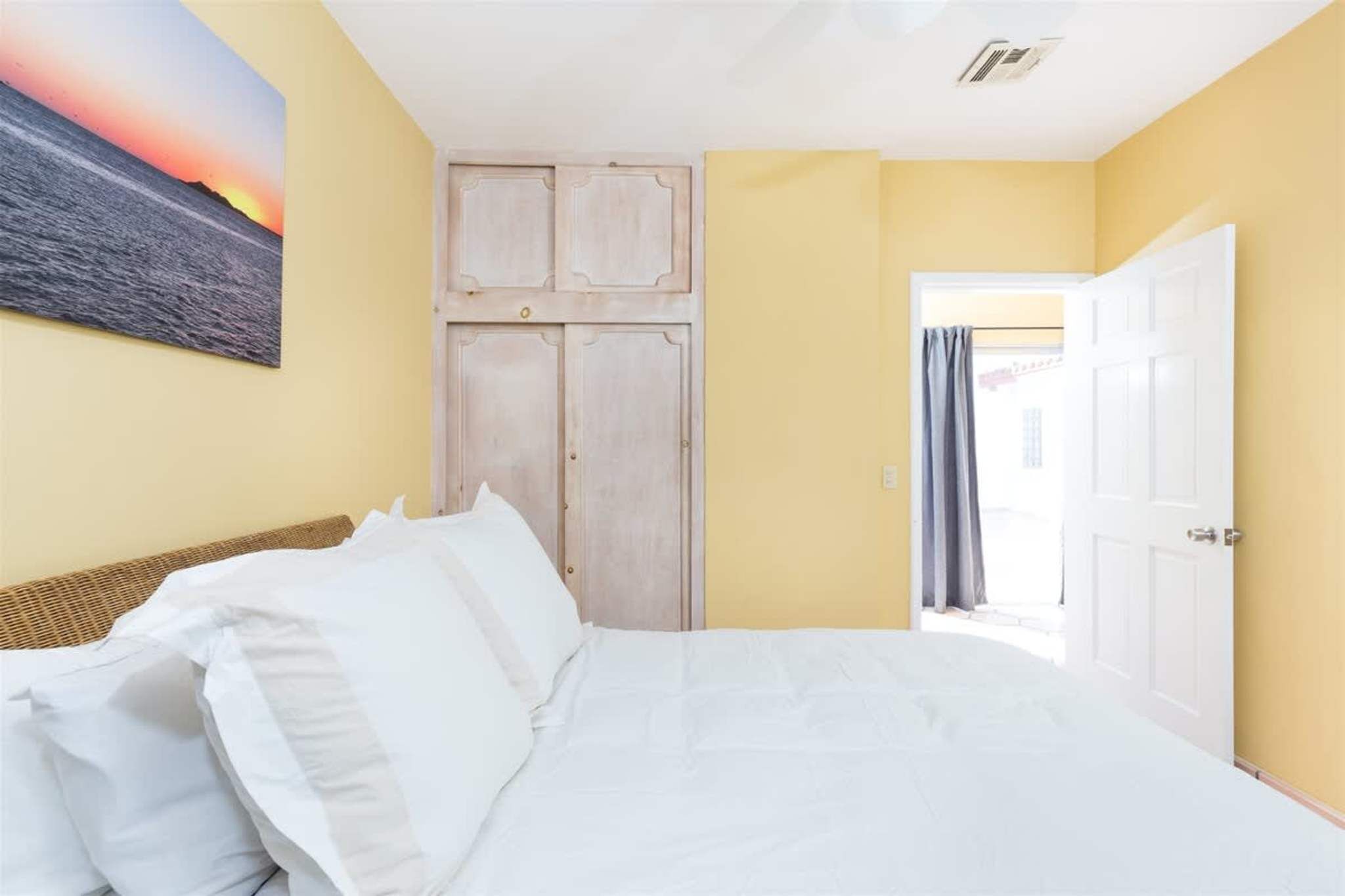 Un dormitorio con una cama, ropa de cama blanca, un cuadro de un atardecer, un armario de madera y una puerta que da a otra habitación.