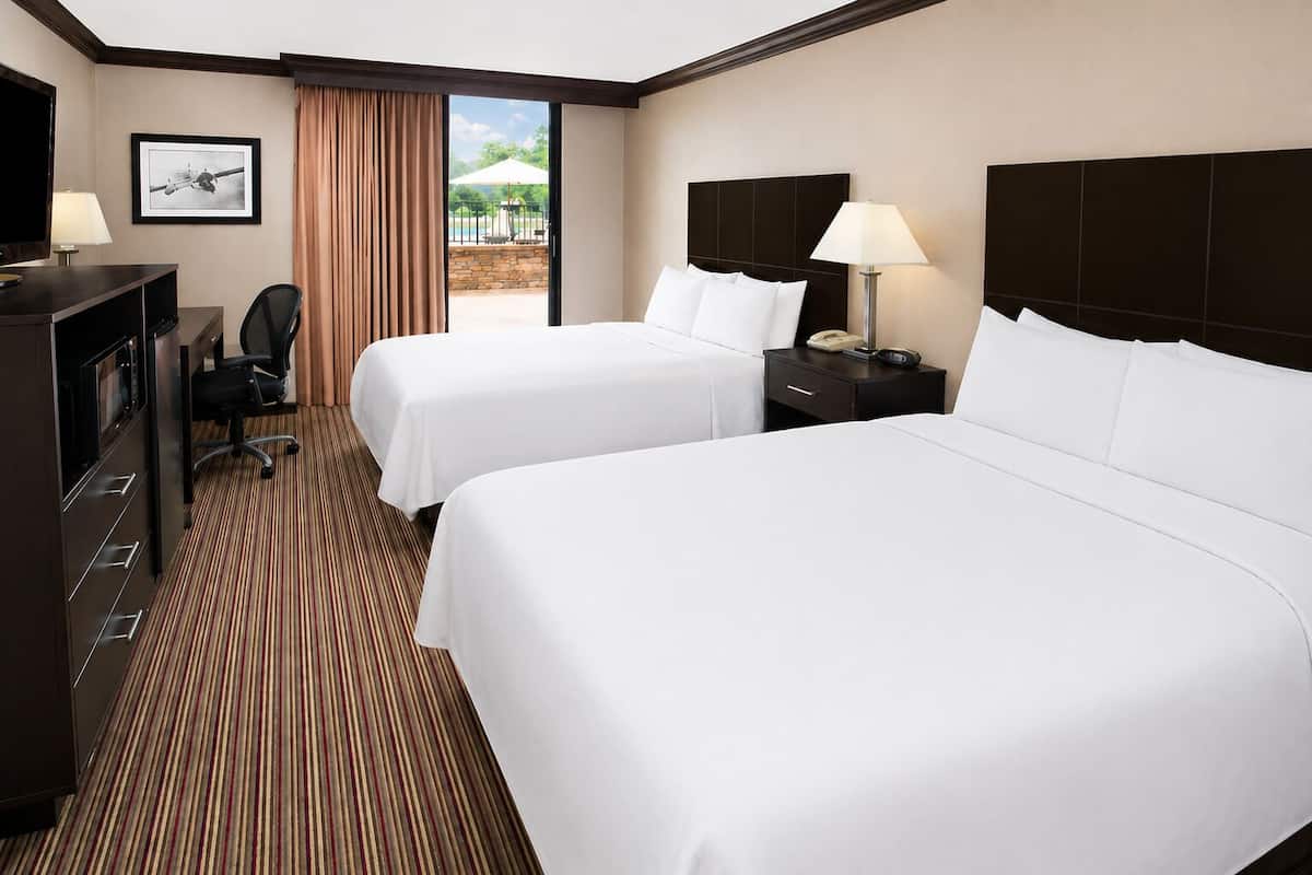 A hotel room with two beds, a desk with a chair, a TV, and a view of an outdoor area.