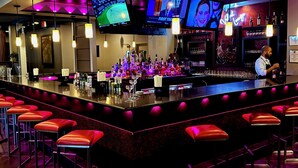 A bar area with multiple screens, a bartender, and red bar stools. - Baymont By Wyndham Schaumburg Chicago Northwest (Schaumburg)