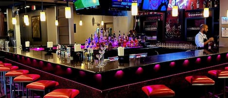 A bar area with multiple screens, a bartender, and red bar stools.
