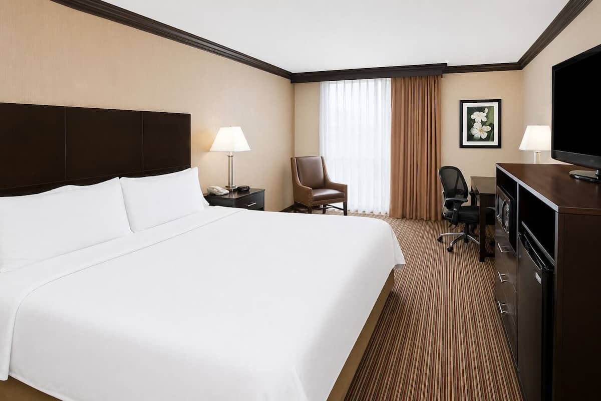 A hotel room with a large bed, a desk with a television, a chair, and a desk lamp.