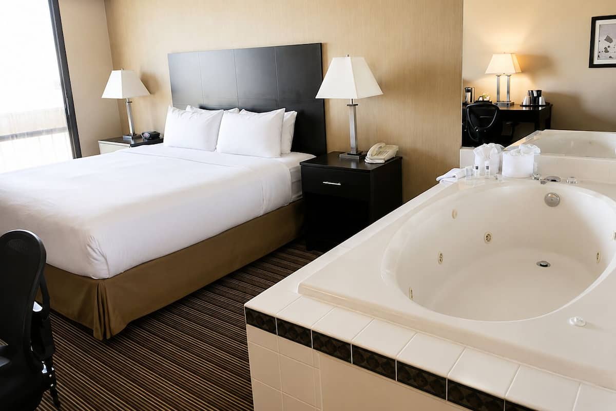 A hotel room with a large bed, two bedside tables with lamps, a black headboard, a large bathtub, and a window.