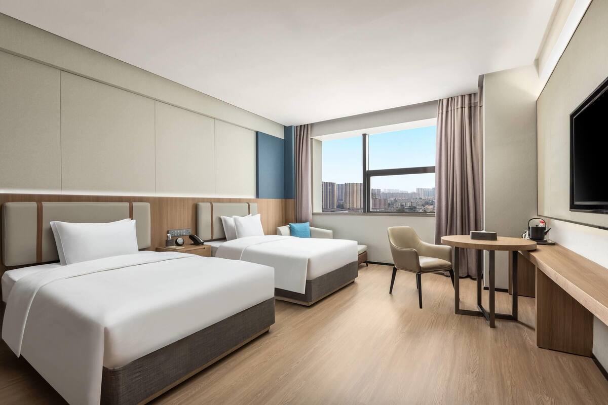 Quarto de hotel com duas camas, uma escrivaninha, uma cadeira e uma janela grande com vista para a cidade.