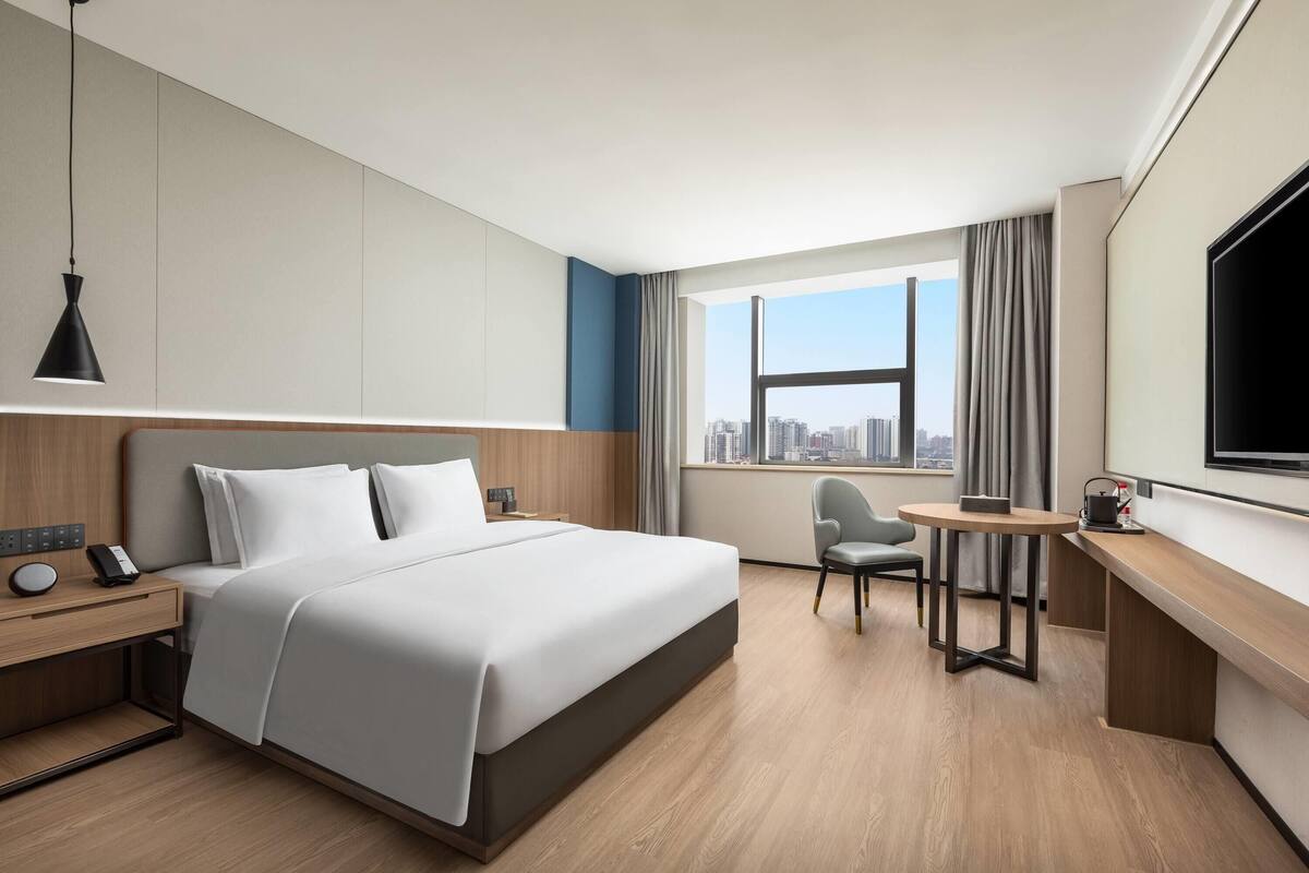Quarto de hotel moderno com cama grande, escrivaninha, cadeira, televisão e vista para a cidade.
