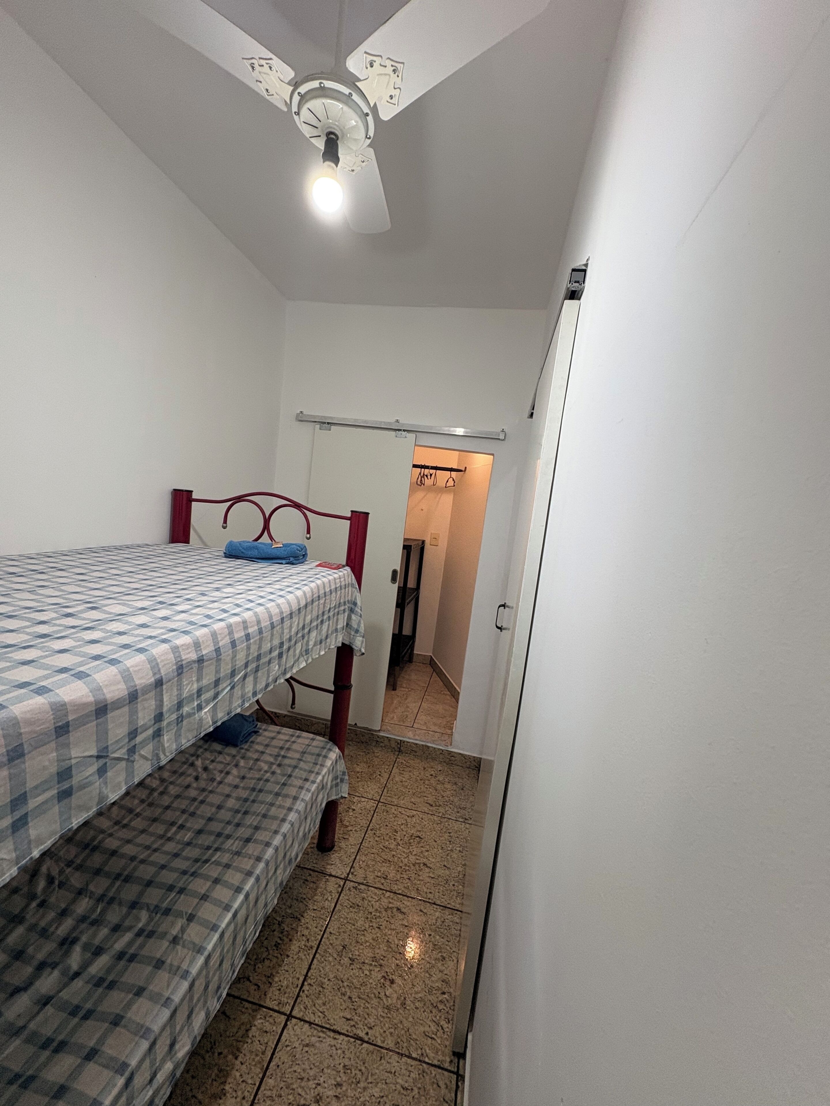 A room with two beds, a ceiling fan, a door leading to another room, and a tiled floor.