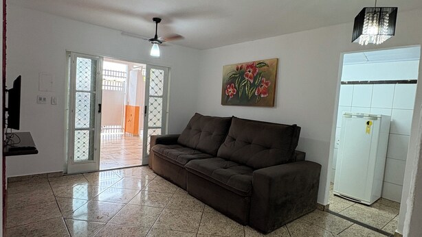 A living room with a brown sofa, a ceiling fan, a painting, and a white refrigerator. - Agrishow house, sleeps 4 / 2 bedrooms / 1 bathroom (Ribeirão Preto)