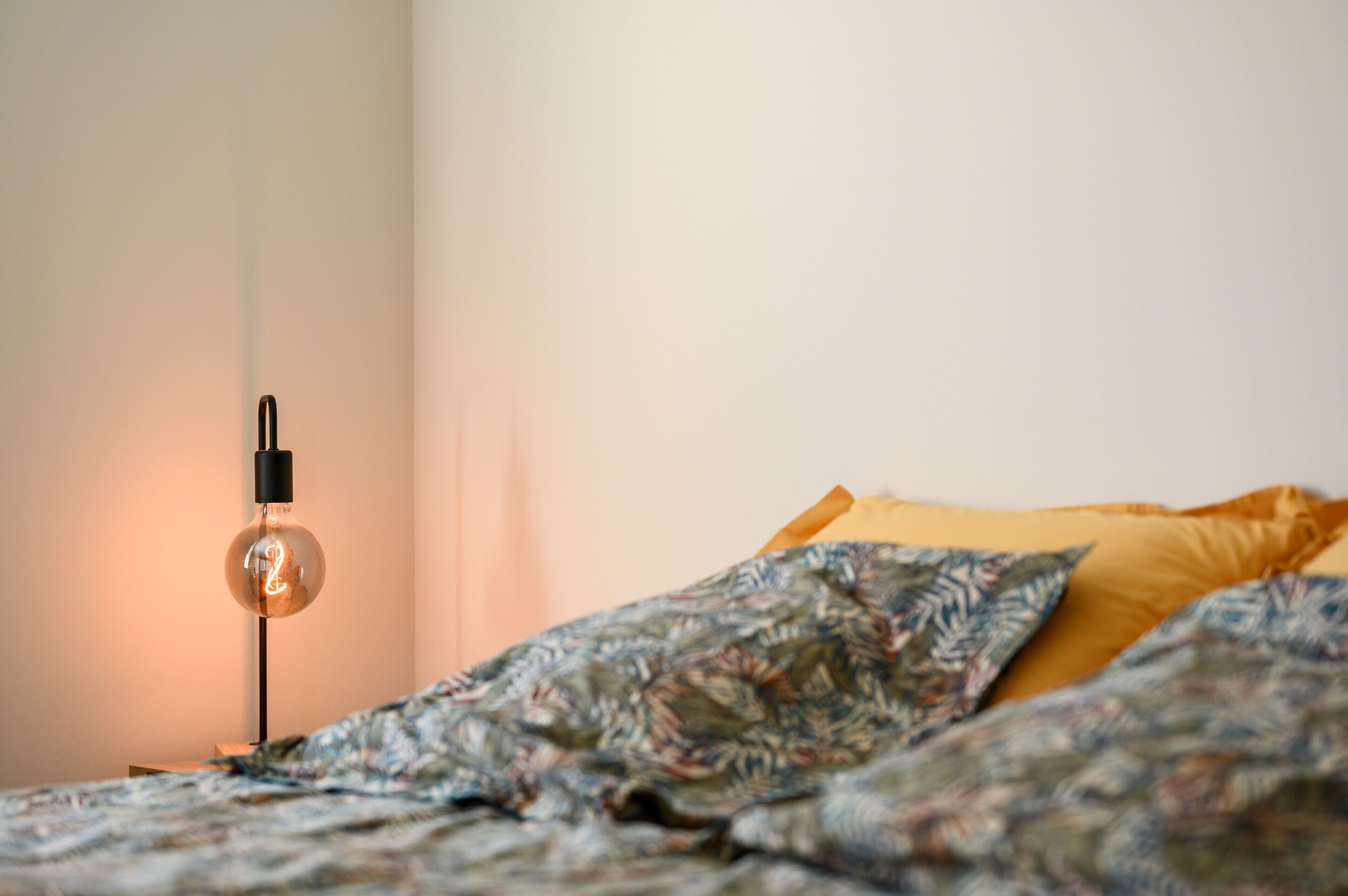 Een bed met een gebedrukte deken, een geel kussen en een wandlamp met een zichtbare gloeilamp.
