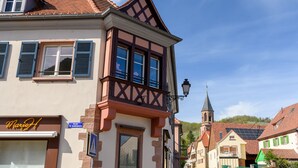 Ein traditionelles europäisches Gebäude mit Satteldach, einem sichtbaren Kirchturm und einem Schild mit der Aufschrift 'Rue de Gonsbach'.