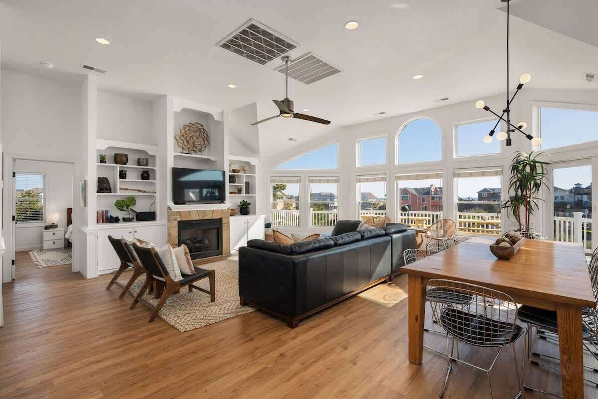 A spacious living room with a fireplace, a black leather sofa, a dining table with chairs, and a large window offering a view of the city.