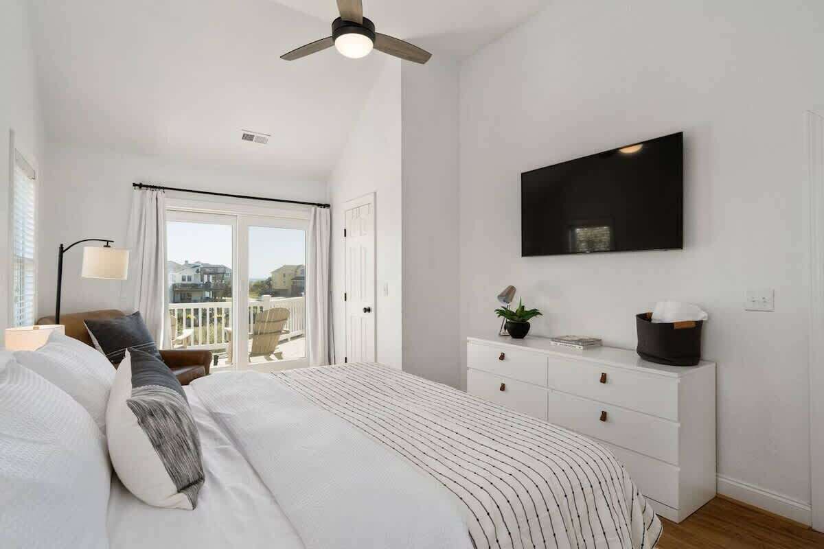A bedroom with a bed, a ceiling fan, a TV mounted on the wall, and a window with curtains.