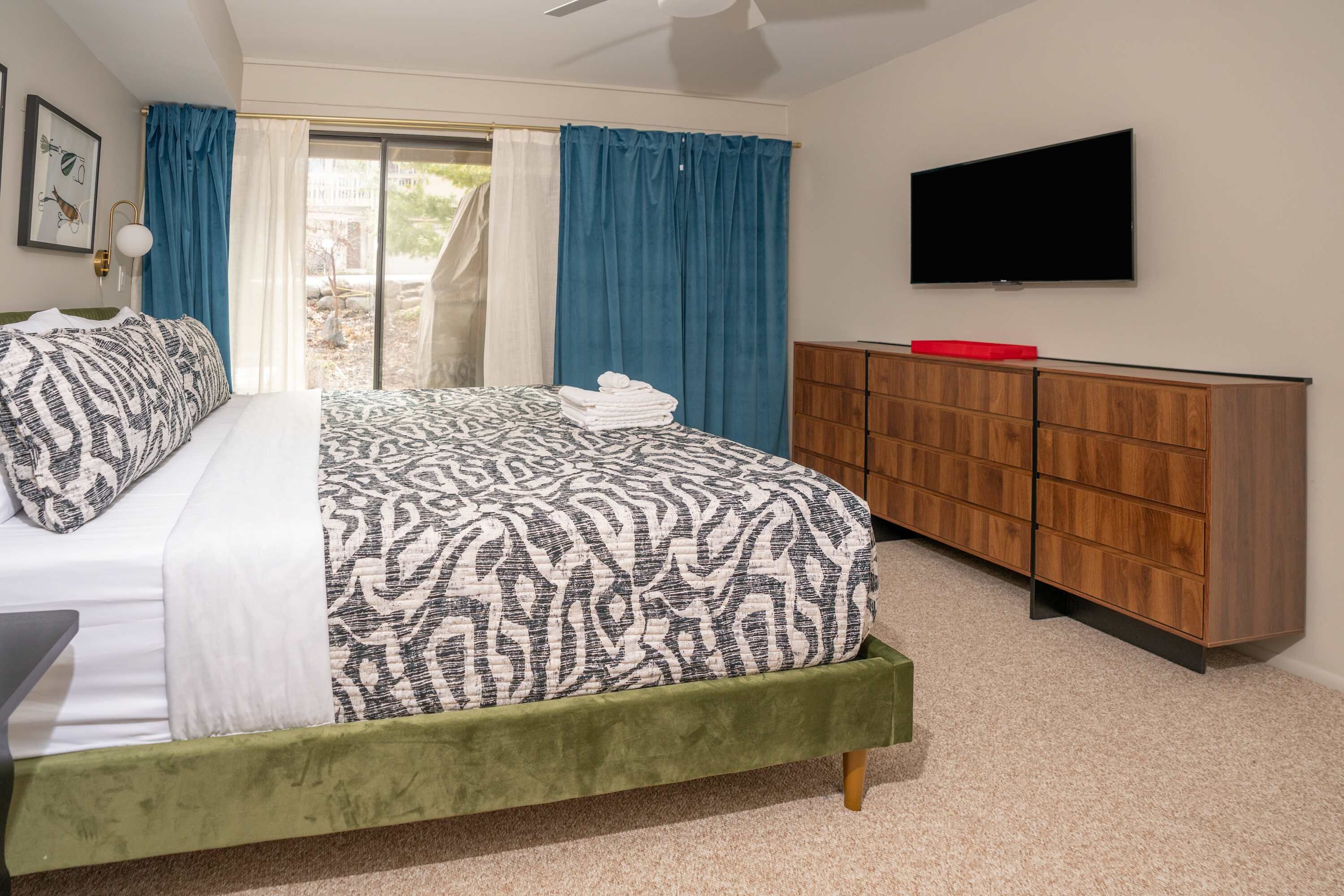 A bedroom with a bed, a TV, a wooden dresser, and a sliding glass door.