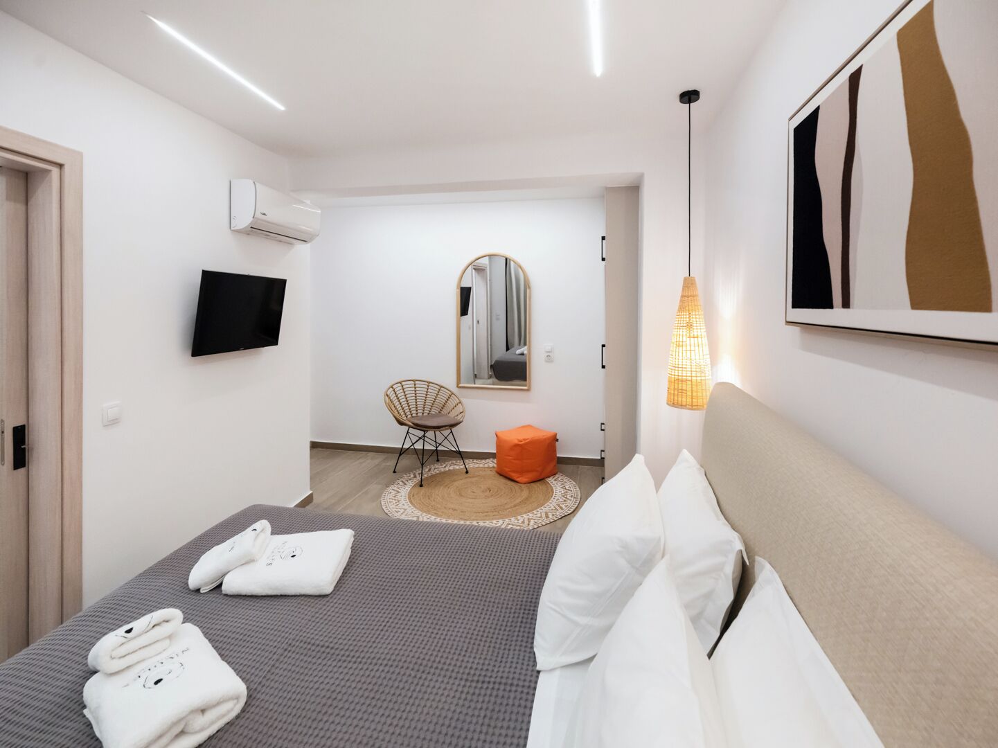 A modern hotel room with a bed, a TV, a mirror, and a hanging light fixture.