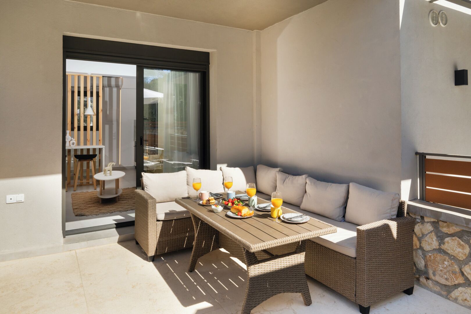 A sunlit patio with wicker furniture, a table set with breakfast, and a view through a glass door.
