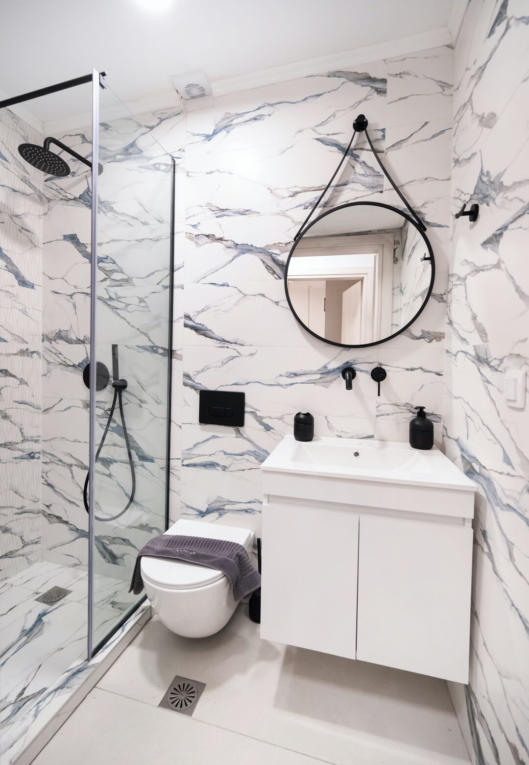 A bathroom with a glass shower enclosure, a white sink with a black faucet, a mirror, and a marble-patterned wall.