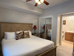 A bedroom with a wooden headboard, a bed with white bedding, a nightstand with a lamp, a mirror, and a door leading to a bathroom. - Coastal Beach Front with Pools, Beach access, Gym (Panama City Beach)