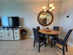 A dining area with a round table, black chairs, a chandelier, a television on a cabinet, and a mirror on the wall. - Coastal Beach Front with Pools, Beach access, Gym (Panama City Beach)