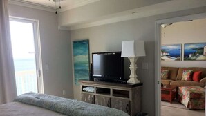 Una habitación de hotel con una cama, un televisor sobre un mueble de madera, un cuadro en la pared y vista al mar a través de una puerta abierta.
