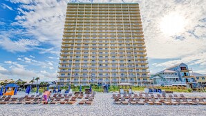 Un hotel alto con varios balcones, una zona de playa con tumbonas y un cielo despejado.