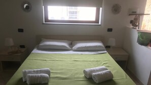 A single bed with a green bedspread, two white pillows, two rolled towels, a nightstand with a lamp, and a small window with a view of buildings. - Cozy studio apartment in rural house with garden 5 km from the sea (Ogliastro Cilento)