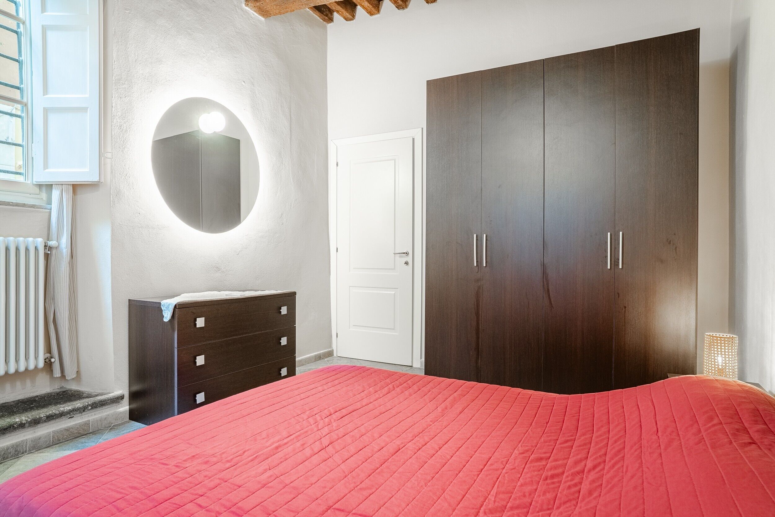 Un dormitorio con una colcha roja, un armario de madera oscura, una puerta blanca y un espejo redondo con luz.