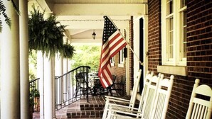 A porch with white rocking chairs, a brick building, and an American flag. - Timeless Elegance at a Historic Kentucky Inn (Versailles)