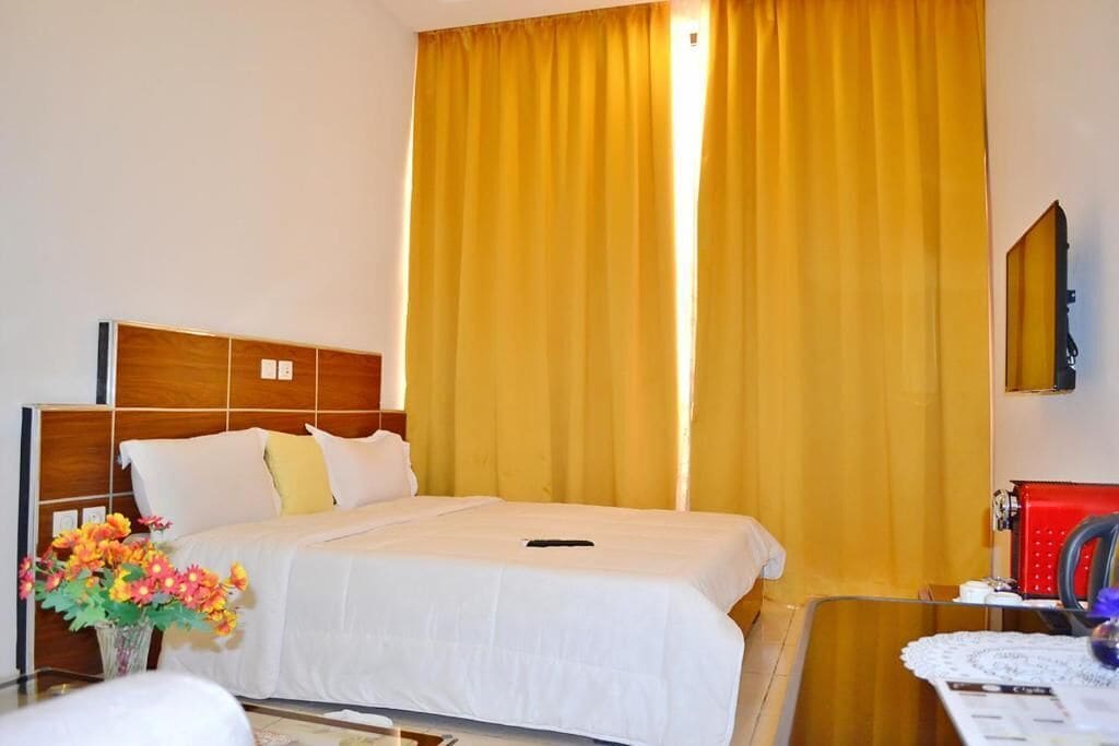 Une chambre d’hôtel avec un lit, une table de chevet, un miroir et une fenêtre avec des rideaux jaunes.