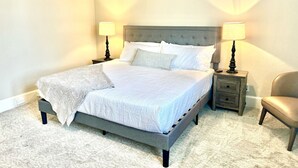 Un lit bien fait, avec une tête de lit grise, deux tables de chevet avec des lampes et une chaise, dans une chambre bien éclairée dont le sol est recouvert d’une moquette.