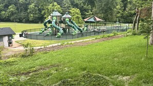 Une aire de jeux avec des toboggans verts et un petit bâtiment, le tout entouré d’herbe et d’arbres.