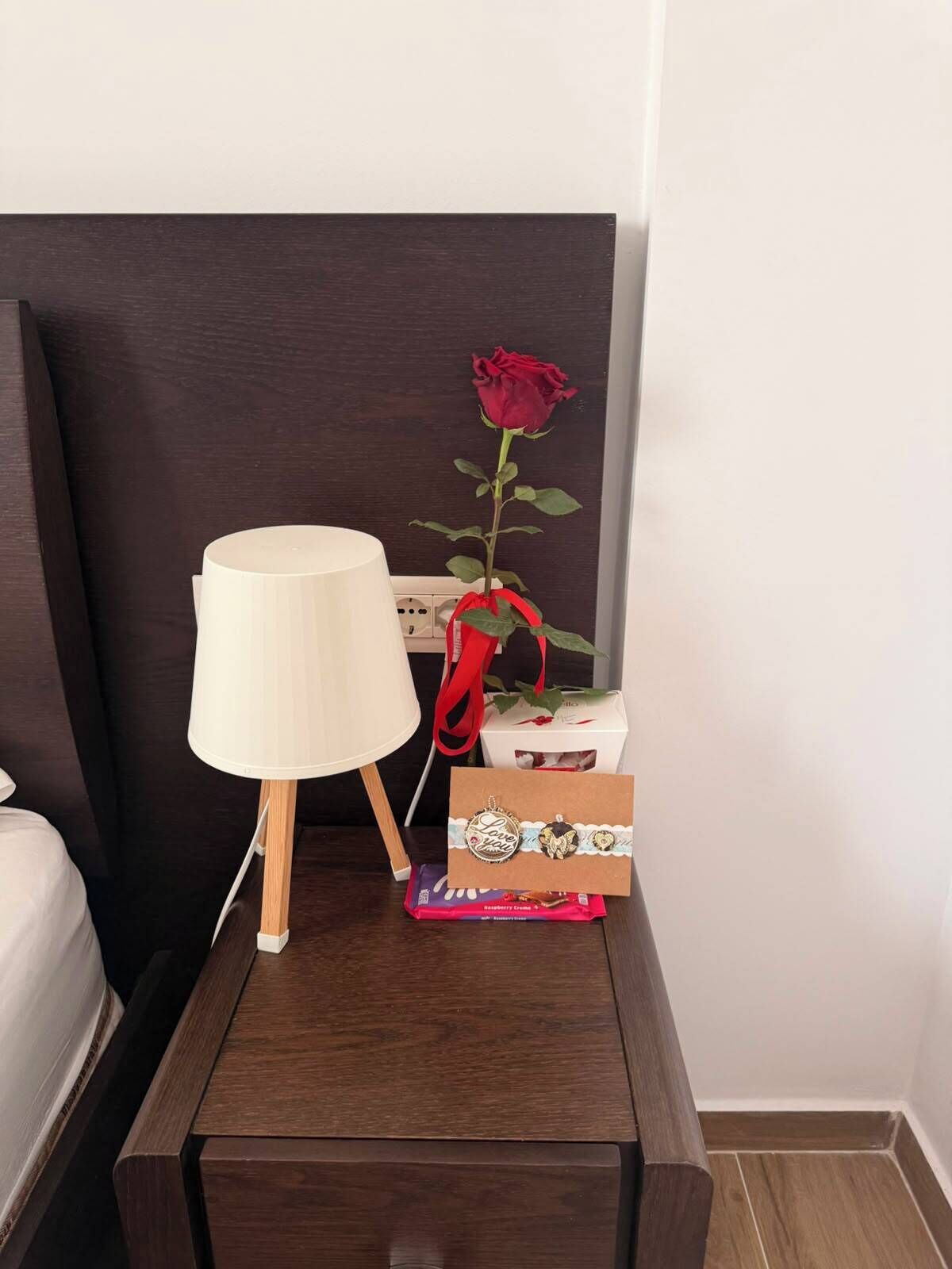 Una mesita de noche con una lámpara, una rosa roja y una caja de regalo.